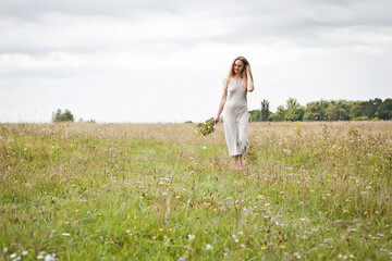 A slender girl in a linen summer dress, with blond long hair and a bouquet of wild flowers in her hands, stands in a clearing during the day.