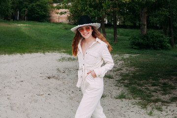 A girl with blond curly hair is dressed in a light overalls and a hat, in sunglasses, stands on the beach and smiles. Hands in pockets. Around the sand against the backdrop of a green park.