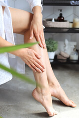 A young woman in a bathrobe touches the soft smooth skin of her legs, applying lotion, doing body...