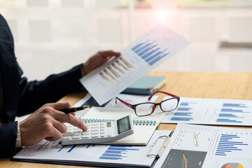 At the accounting desk, an accountant presses a calculator to analyze investment charts to generate...