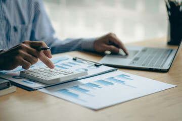 At the accounting desk, an accountant presses a calculator to analyze investment charts to generate monthly financial reports or company profits.