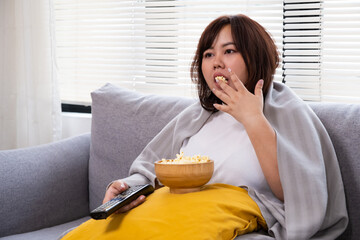 Young Asian oversize women gain weight while eating donut, bread and snack while watching television. Junk food that can cause obesity and unhealthy for human body. - obrazy, fototapety, plakaty
