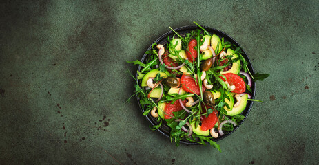 Fototapeta na wymiar Healthy vegan salad with arugula, avocado, juicy grapefruit, cashews and dressing with olive oil, honey and wine vinegar. Green rustic kitchen table, top view, copy space