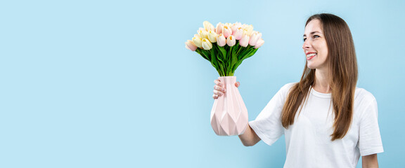 Smiling young woman looking at tulips in vase on blue background. Banner