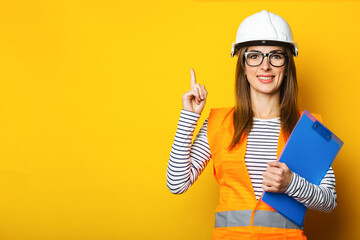 Young woman with a smile in a vest and hard hat holds a clipboard on a yellow background....