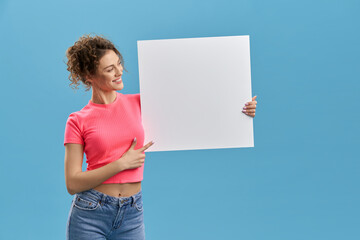 Cute girl in outfit smiling, holding big sheet of white paper in studio, copy space. Portrait view...