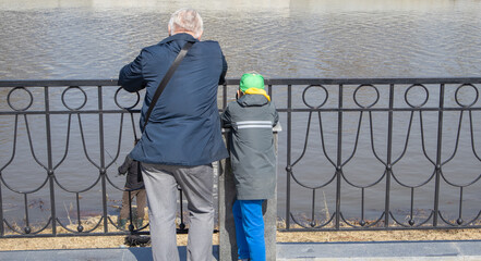 A gray-haired grandfather and his grandson are standing on the river embankment and leaning on metal handrails, looking at a fisherman on a sunny spring day