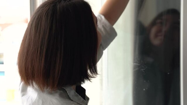 Beautiful smiling young woman cleaning and wiping window with spray bottle and rag. housework and housekeeping concept . face reflection in glass. 4k footage.