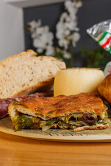 Pizza cotta al forno ripiena di scarole, olive e acciughe con sullo sfondo del pane caldo e una forma di formaggio fresco