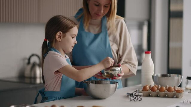 little girl is helping to her mom in kitchen, mother and daughter are cooking together, adding sugar