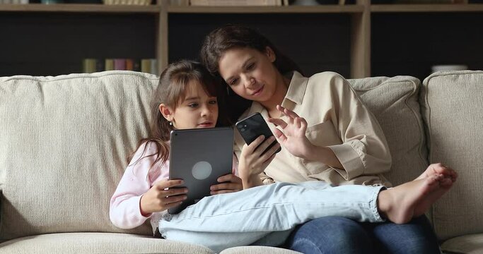 Young mom holds smartphone her daughter use digital tablet spend leisure at home with wireless gadgets, have fun, discuss new cool app. Young gen and modern tech overuse, on-line games users concept