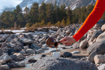 Girl's hand holding coffee pot pours coffee into porcelain cup on big stone against mountain river
