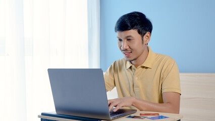 Handsome Asian man wearing a yellow shirt pays for online shopping on his laptop computer. The young man looked happy after completing his credit card payment. online shopping