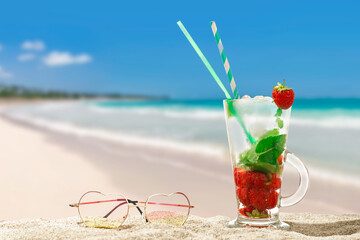 Glass of fresh mojito cocktail and sunglasses on beach sand