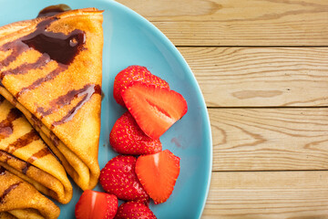 Delicious thin pancakes with fresh strawberries and chocolate on a blue plate. Wooden background. top view