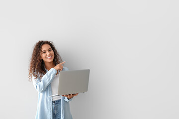 Beautiful African-American woman with laptop pointing at something on light background - obrazy, fototapety, plakaty