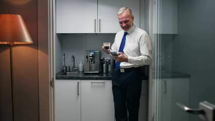 Naklejka na ściany i meble Senior executive drink coffee and use smartphone standing in corporate kitchen