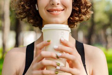 Young redhead girl smiling happy wearing sports bra standing on city park, outdoors holding a takeaway coffee mug front of chest and closed eyes. Coffee lover and outdoor sport concepts.