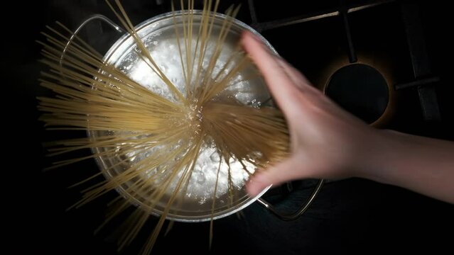 Hand of chef throwing spaghetti pasta into boiling water in silver pot on kitchen stove, top view. Italian cuisine, cooking, food preparation.