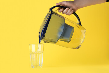 Woman pouring purified water into glass from filter jug on yellow background