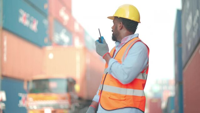 African American cargo container worker as foreman or technician use walkie talkie to comunicate with his co-worker in workplace with truck run pass him and he smile to greeting.