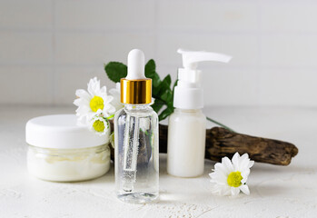 Fototapeta na wymiar Facial skin care fluid and skin cream on the white table. A twig with chamomile flowers in the background. Light brick background