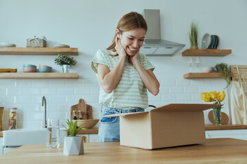 Surprised young woman unpacking box while standing at the domestic kitchen