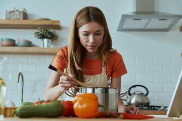 Confident young woman cooking soup while standing at the domestic kitchen