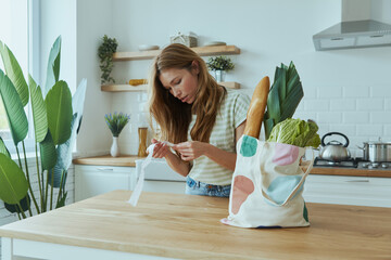 Concentrated young woman checking receipts while standing at the domestic kitchen
