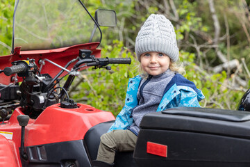Portrait of a smiling four year old boy outdoors wearing a grey wooly hat and blue coat. Sitting on a rural quad bike with blurry forest background.