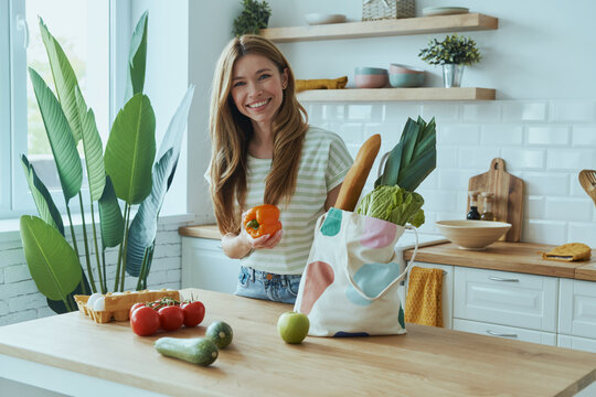 Beautiful young woman unpacking the bag with healthy food while standing at the domestic kitchen - Powered by Adobe