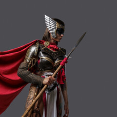 Shot of female barbarian holding spear dressed in steel armor with helmet against grey background.