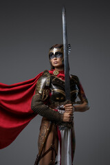Fototapeta premium Shot of female barbarian holding sword dressed in steel armor with helmet against grey background.