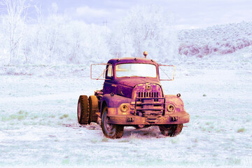 Abandoned truck sits in an empty field with white washed hills and trees in the background. The vintage truck is tinted orange, purple and yellow and is empty but still has its tires.