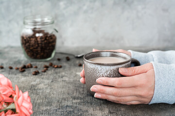 Latte in a ceramic cup in female hands, a jar of coffee beans and roses on the table.