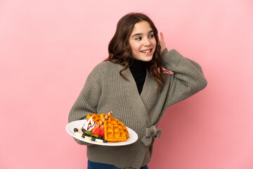 Little girl holding waffles isolated on pink background listening to something by putting hand on the ear