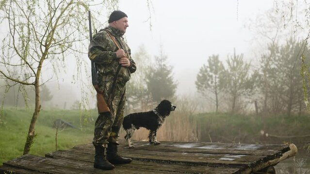 A hunter with a gun stands on a lake bridge with a dog and looks around