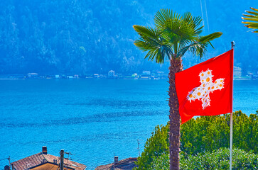 The flag of Switzerland and palm tree in front of Lake Lugano, Morcote, Switzerland