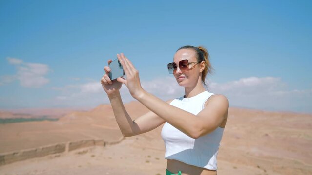 Tourist woman using cell phone for shooting, standing in desert. Action. Young female taking picture of desert on her Iphone.