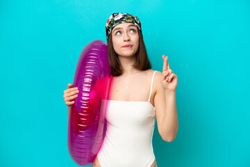 Young Ukrainian woman holding air mattress isolated on blue background with fingers crossing and...