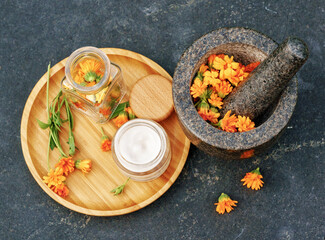 Calendula Cream for body care in glass jar and fresh orange calendula flowers on a wooden plate and a granite mortar on black table. Concept: spa, aromatherapy, Medical Dermatology. Flat lay - obrazy, fototapety, plakaty
