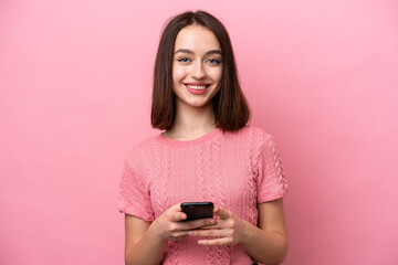 Young Ukrainian woman isolated on pink background looking at the camera and smiling while using the mobile
