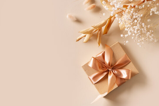Gift box with golden ribbon and dry grass and flowers on beige background