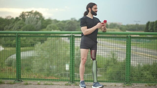 long black hair caucasian bearded man disabled artificial leg smiling and leaning against railing bridge holding mobile phone texting message after workout