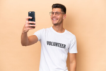 Young volunteer caucasian man isolated on beige background making a selfie