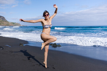 A young woman has fun jumping on the shore of a black sand beach.