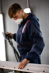 Mechanic in protective mask and suit holding sander near car part in auto service station 