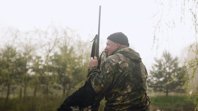 A hunter with a weapon is sitting near a misty lake next to his friend the hunting dog, the dog licks his face showing his love which makes his master hunter smile