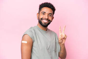 Young Brazilian man wearing a band aid isolated on pink background smiling and showing victory sign