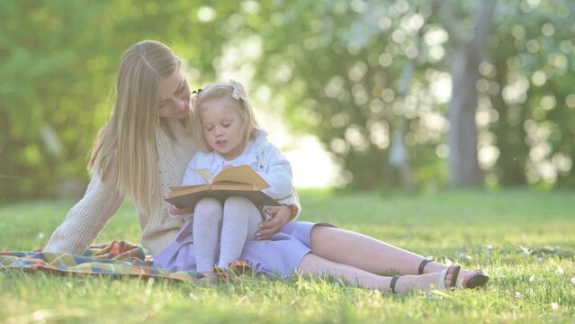 Cheerful young mum and baby reading a book outdoor beautiful field of sunshine and spring grass while on a summer holiday. Family outdoor lifestyle.